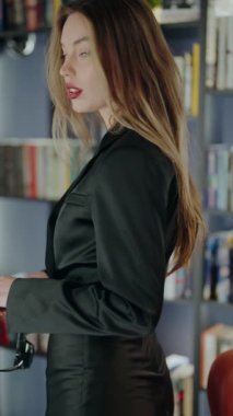 Siyah elbise ve güneş gözlüğü takan şık bir kadın modern bir kütüphanede güven içinde poz veriyor. Dekor sofistike ve zariftir. Siyah elbiseli, güneş gözlüklü şık bir kadın. Dikey