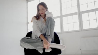 Gündelik beyaz gömlek ve kot giymiş, parlak bir pencerenin yanındaki modern bir sandalyeye tünemiş genç bir kadının huzurlu portresi. Günlük kıyafetli genç bir kadın Düşünceli bir şekilde sandalyede oturuyor.