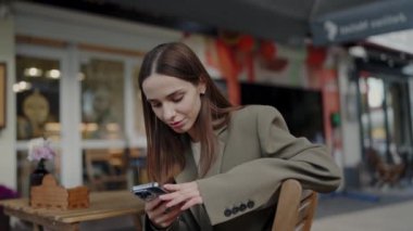 Açık Kafe Masası 'nda akıllı telefon kullanan genç bir kadın. Dışardaki bir kafede oturmuş şık bir palto giymiş akıllı telefonunu karıştıran odaklanmış genç bir kadın..