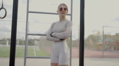 Kendine güvenen sporcu kıyafetli genç bir bayan sporcu açık hava fitness parkında kolları bağlı duruyor ve kararlılık ve hazırlık gösteriyor. Kendinden emin genç sporcu güçlü duruyor.
