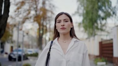 Zarif genç bir kadın, şık beyaz bir gömlek içinde, düşünceli ve sakin bir ifadeyle, güvenli bir şekilde şehir caddesinde yürüyor. Beyaz Gömlekli Genç Kadın Şehir Caddesinde Yürüyor