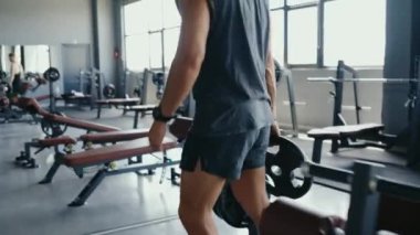 Spor salonundaki halterlere ağırlık ekleyen bir adamın, hazırlık ve zindeliği vurguladığı yakın plan çekimi. Görüntü spor salonunun atmosferini yakalıyor ve güç antrenmanına odaklanıyor. Halteri Hazırlayan Adam