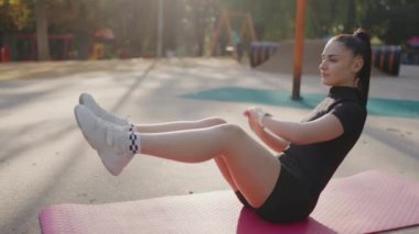 Mat 'in üzerinde esneme egzersizi yapan genç kadın. Odaklanmış genç bir kadın parkta pembe bir minder üzerinde karın egzersizleri yapıyor, güneşli bir açık hava fitness seansının tadını çıkarıyor..
