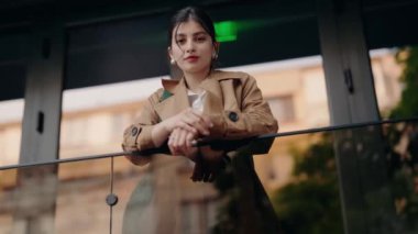 Trençkotlu, kendine güvenen genç bir kadının portresi, modern mimariyle şehir zeminine karşı bir balkonda duruyor. Trençkotlu Balkonda Duran Kendinden emin Genç Kadın