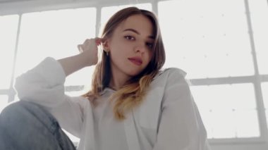 Nazik bakışlı genç bir kadın pencerenin yanında poz veriyor, iyi aydınlatılmış bir odada huzurlu ve düşünceli bir aura yayıyor. Beyaz Gömlekli Genç Kadın Düşünceli Poz veriyor Pencere kenarında