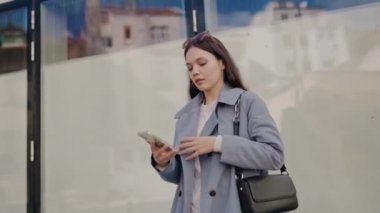 Şehirde cam bir binanın yanında yürürken akıllı telefonunu kullanarak şık bir trençkotla genç bir kadına odaklanmış. Modern yaşam tarzı ve bağlantıyı yakalar.