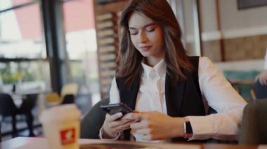 İş kıyafeti giymiş profesyonel bir genç kadın bir şehir kafesinde akıllı telefonuna takılıp kalmış. Odaklanmış Genç İş Kadını Bir Kafe Ayarında Akıllı Telefon Kullanıyor
