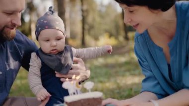Ebeveynler bebeklerinin ilk doğum gününü güneşli bir parkta pastayla kutlarken neşeli bir aile anı. Aşk, mutluluk ve açık havada paylaşılan özel bir durum..
