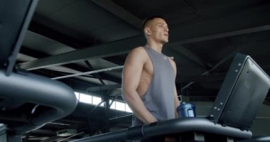 Modern Spor Salonu 'nda Koşu bandında Egzersiz Yapan Odaklanmış Adam. Gri kolsuz tişörtlü fit bir genç, geniş, aydınlık bir spor salonunda elinde su şişesi tutarken koşu bandı kullanıyor..