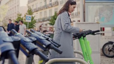 Gri montlu genç bir kadın kalabalık bir şehir caddesindeki liman istasyonundan elektrikli scooter seçiyor. Yoğun Şehir Ortamı 'nda Elektrikli Scooter Kiralayan Genç Kadın