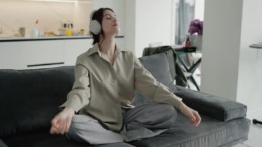 Bir kadın kanepede bacak bacak bacak üstüne atarak oturur, kulaklık takarak meditasyon yapar, rahat bir ev ortamında rahatlık ve farkındalığı somutlaştırır. Evde kulaklıklarla dinlenen, müzikle eğlenen bir kadın.