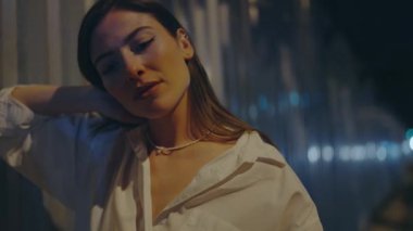 Beyaz gömlekli düşünceli genç bir kadın geceleri tek başına, yumuşak şehir ışıklarının arasında, yalnızlık ve içgözlem temalarını yansıtıyor. Kentsel Ayarlarda Düşünceli Genç Kadın