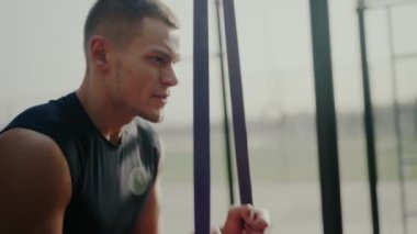 Siyah kolsuz tişörtlü bir erkek sporcu arka planda spor yüzükleriyle zihinsel olarak açık bir spor salonunda hazırlanırken görülüyor. Bağlılığı ve uygunluğu temsil ediyor. Açık havada Odaklanmış Atletizm Eğitimi