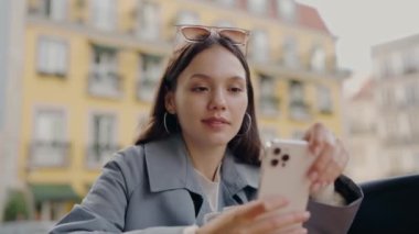 Genç bir kadın dışarıda oturuyor, odaklanmış bir bakışla akıllı telefonuna bakıyor. Parlak, renkli binalar canlı bir zemin oluşturur. Sunny City Caddesinde Akıllı Telefon Kullanan Genç Kadın