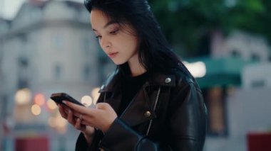 Siyah deri ceketli genç bir kadın şehir ortamında dışarıda dikilirken dikkatle akıllı telefonuna odaklanıyor. Deri ceketli genç bayan dışarıda akıllı telefon kullanıyor.