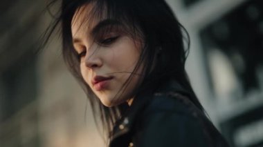 Koyu renk saçlı ve dalgın suratlı genç bir kadının yakın plan fotoğrafı. Arka planı bulanık, açık bir şehir ortamında duruyor. Açık havada rüzgarlı saçları olan düşünceli bir kadının portresi.