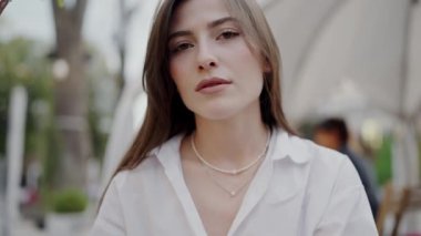 Sakin bir ifadeyle şehirde beyaz gömlek giyen genç bir kadının yakın plan portresi. Bulanık şehir arkaplanı ortam katıyor. Meşgul Şehir Caddesinde Beyaz Gömlekli Genç Kadın Pozu