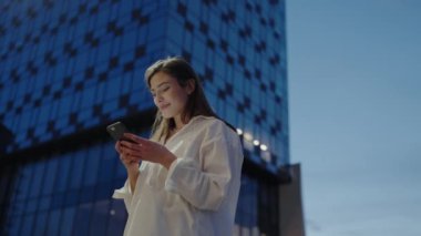 Gündelik beyaz gömlekli genç bir kadın, alacakaranlıkta modern cam bir binanın önünde duran bir akıllı telefon kullanıyor. Sahne, yoğun ama huzurlu bir şehir hayatını yansıtıyor..