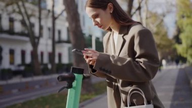 Şık montlu genç bir kadın, güneşli bir şehir caddesinde elektrikli scooterıyla dururken akıllı telefonunu kullanıyor, modern şehir hareketliliğini ve bağlantılılığını somutlaştırıyor..