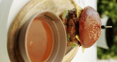 Peynirli, sebzeli ve soslu sulu sığır etli burger bulanık arka planda masada yavaşça döner. Biftek Burger. Biftek burgeri bir restoranın terasında servis edilir. Hikayeler için dikey video