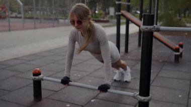 A focused young woman in sunglasses trains on parallel bars at a park, demonstrating determination and fitness. Young Woman Exercising on Outdoor Parallel Bars in the Park