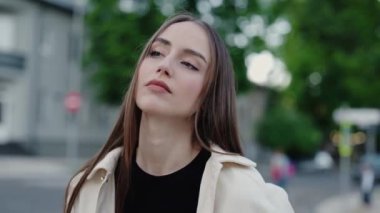 Uzun saçlı ve tarafsız ifadeli genç bir kadın şehir ortamında, derin düşüncelere dalmış bir şekilde dışarıda duruyor. Arka planda ağaçların ve binaların olduğu bir cadde var..
