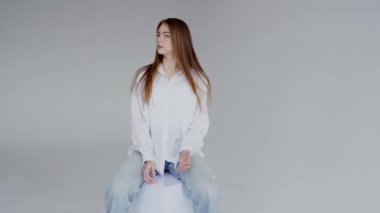 Beyaz gömlek ve kot pantolon giyen mutlu genç bir kadının portresi gri bir stüdyo arkaplanına oturup gülümsüyor. Gri Arkaplanda Beyaz Gömlekli Gülümseyen Genç Kadın pozu