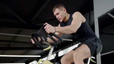 Odaklanmış genç bir adam, modern bir spor salonundaki sabit bir bisiklet makinesine pedal çevirirken, yoğunluğunu ve uygunluğunu gösterir. Azimli Genç Adam Spor Salonunda Sıkı Çalışıyor
