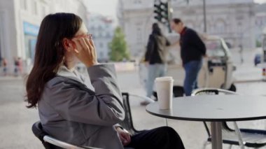 İş kıyafeti giymiş bir kadın, arka planda hareketli bir şehir ortamı ve masanın üzerinde kahve fincanı olan bir kaldırım kafesinde otururken telefonuyla sohbet ediyor..