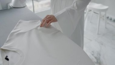Bir kadın, temiz, minimalist bir ortamda, ev işlerini gösteren bir ütü masasında beyaz bir gömlek ütülüyor. Parlak Modern Daire 'de Giysileri Ütüleyen Kadın