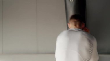 Spor salonunda kum torbasıyla egzersiz yapan, uzun kollu beyaz bir atletik bluz giyen odaklanmış genç bir adam. Spor Salonunda Ağır Kum Torbasıyla Genç Atletizm Eğitimi