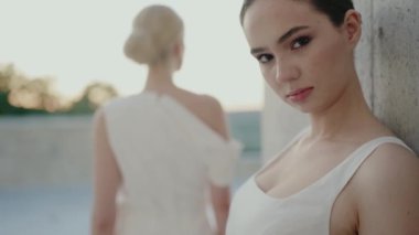 Beyaz elbiseli iki zarif kadın, biri kameraya bakıyor ve diğeri arkasını dönüyor, açık hava ortamında güven ve sükunet yayıyorlar. Beyaz Elbiseli Zarif Kadınlar