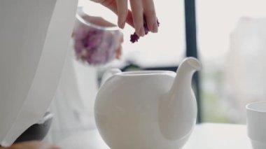Beyaz çaydanlığa kurutulmuş bitkisel çiçekler ekleyip evde bitki çayı karışımı hazırlayan birinin yakın çekimi. Rahat ve sakin bir sabah rutini. Evde Beyaz Çaydanlıkta Elle Bitki Çayı Hazırlama