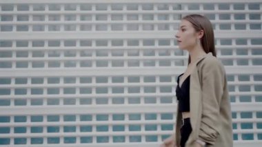 Tarz sahibi bej ceketli genç bir kadın grafiksel ve modern bir binanın önünde güvenle duruyor. İfadesi derin derin düşünüyor ve sakin bir şehir havası yayıyor..