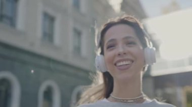 Şehrin arka planında neşeli bir genç kadın, beyaz kulaklıklarıyla müzik dinlerken gülümsüyor, günlük kıyafetlerini giyiyor. Kentsel Ayarlar 'da Kulaklıkla Müzikten Hoşlanan Genç Kadın