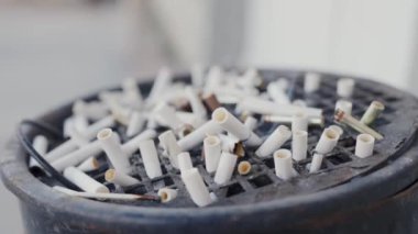 Sigara izmaritlerinin yakın çekimi. Kül dolu büyük metal bir kül tablasında sigara izmaritleri yakmak. Çöpe atılmış, filtrelenmiş sigara izmaritlerinin yakın görüntüsü.