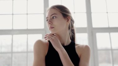 Kolsuz bir bluz giyen düşünceli genç bir kadının portresi. Yumuşak ışıklandırılmış bir odada geniş pencerelerin yanında düşünceli bir şekilde poz veriyor. Düşünceli Genç Kadın Büyük Parlak Pencerelerin Yakınında Poz Veriyor