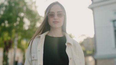 Güneşli bir parkta, güneş gözlüğü takan ve sıradan bir ceket giyen gülümseyen bir kadının portresi. Kendinden emin ve rahat görünüyor. Dışarıda keyifli bir gün geçiriyor. Güneş gözlüklü Gülümseyen Kadın
