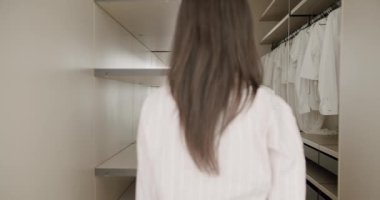 Uzun saçlı bir kadın boş rafları ve asılı kıyafetleriyle modern bir dolaba giriyor. Minimalist ve organize bir yer. Dolapta duran, rafları ve asılı giysileri olan kadın.