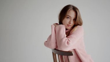 Düşünceli, pembe bir kazağa sarılı genç bir kadın, tahtadan bir sandalyenin arkasına yaslanmış, sakin ve düşünceli bir tavır sergiliyor. Pembe Süveterli Genç Kadın Tahta Sandalyeye yaslanıyor