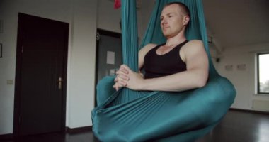 Sakin erkeğin yeşil hamakta oturduğu ve sabahları stüdyoda yoga yaparken kapalı gözlerle meditasyon yaptığı görüntüyü büyüt. Adam hava hamağında meditasyon yapıyor..