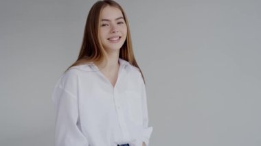 Yumuşak gri bir arka planda beyaz bir bluz giymiş, nazikçe gülümseyen genç bir kadının portresi. Yaşam tarzı ve moda kullanımı için mükemmel. Rahat giyimli genç bir kadın. Taburede Serenely oturuyor.