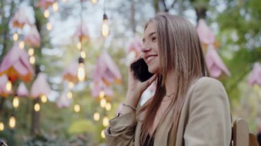 Parkta oturmuş ışıkları ve üçgen bayraklarla süslenmiş telefonuyla gülen neşeli bir genç kadın. Hoş Bir Telefon Görüşmesi Yapan Genç Kadın