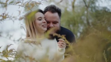 Gün batımının sıcacık ışığında yumuşak yapraklarla çerçevelenmiş, sevgiyle gülümseyen genç bir çiftin romantik bir sahnesi. Günbatımında Çiçek açan bir bahçede kucaklaşan romantik çift