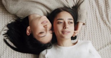 Low angle handheld shot of young lesbian couple smiling and touching hands gently while sitting on sofa against window in light living room at home. Lifestyle and relationships. LGBT concept