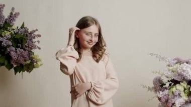Güzel sarışın 13 yıllık bej elbiseli ve gözlüklü genç kız mutlu ve gülümsüyor ve stüdyoda doğal çiçeklerle beyaz bir arka planda kameraya bakıyor. Güzel genç kız gülümsüyor..