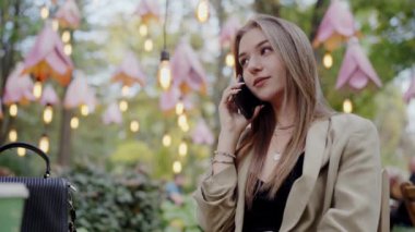 Süslü, aydınlık bir kafede oturan çekici genç bir kadın cep telefonuyla sohbet ediyor. Mekan samimi ve davetkar bir atmosfer yayıyor. Genç Kadın Dışarıdaki Kafe 'yi arıyor.