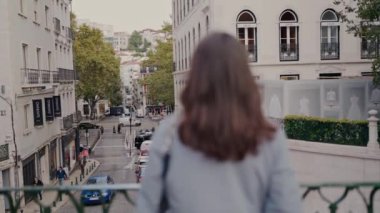 Köprüde dikilen genç bir kadın ağaçlar ve tarihi binalarla dolu hareketli bir caddeye bakıyor. Bir şehir keşfi anı yakalandı. Yoğun Şehir Caddesine Bakan Genç Kadın