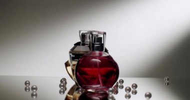 Kırmızı bir sıvıyla donatılmış sofistike bir parfüm şişesi yansıtıcı bir yüzeydeki dağınık incilerin arasında zarif bir şekilde duruyor ve lüks ve büyüleyici bir ortam yaratıyor. Zarif parfüm şişesi ve inciler.