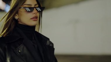 Deri ceket giyen genç bir kadın şık marka güneş gözlükleriyle fotoğraf çekimi için poz veriyor. Moda estetiği modeli. Deri ceketli ve gözlüklü çekici bir kadın merdivenlerde bekliyor..